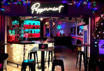 Interior of Peppermint Gay Bar in Gran Canaria, featuring festive decor, bar with colorful lighting, tables, and stools. Gay travel.