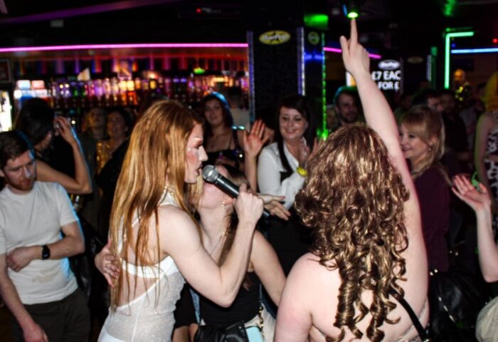 Manchester gay bar scene: Drag queen performs karaoke in a lively, crowded bar. People cheering, enjoying the vibrant nightlife.