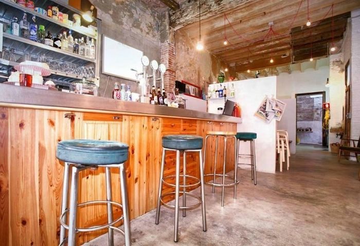 Interior of Bar Rufián, a gay bar in Barcelona. Stools line the wooden bar. Shelves of liquor bottles. Rustic design with exposed brick.