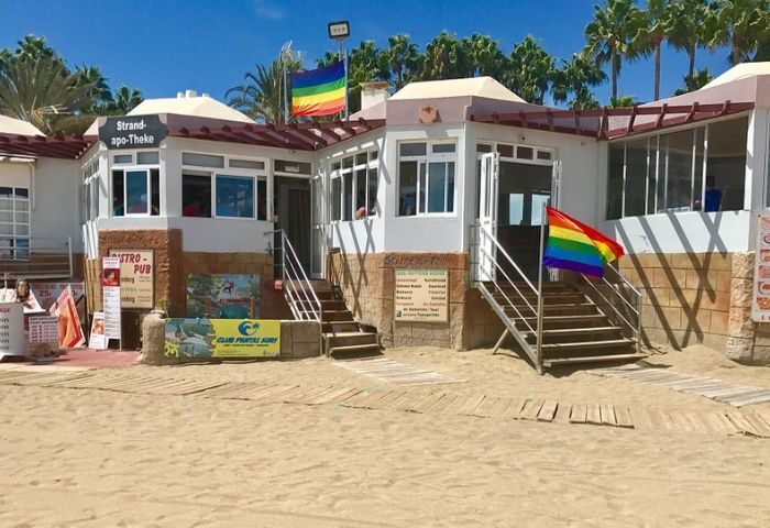 Strand-Apo-Theke gay bar in Gran Canaria. Beachfront bar with rainbow flags. Popular gay travel destination. Palm trees in background.