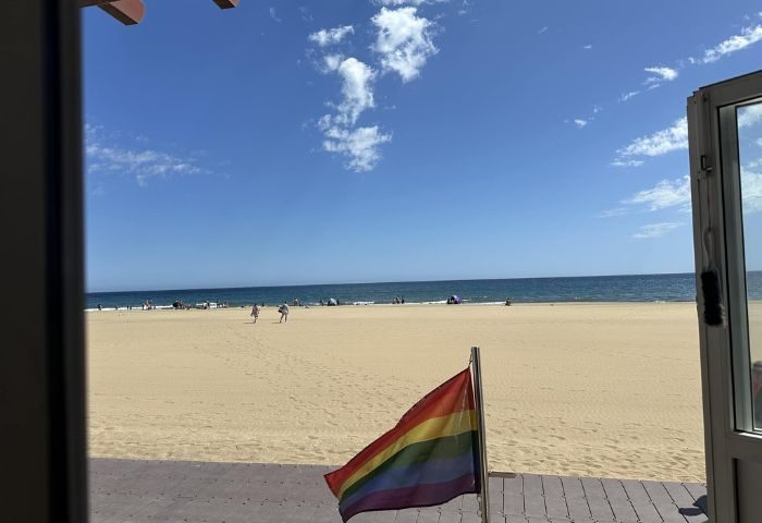 Gran Canaria gay bar view: Beach scene with rainbow flag. Sunny day, blue sky, ocean. Gay travel destination. Apo-Theke bar.