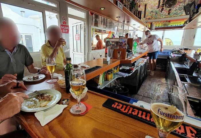 Gran Canaria gay bar: Patrons enjoy food and drinks at a beachside bar. A bartender wearing a mask is visible. Rainbow flag.
