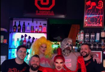 Bar TR3S Lisboa: Drag queens and gay men pose at the gay bar. Neon sign with male figure and Youareabottom visible. Travel advice.