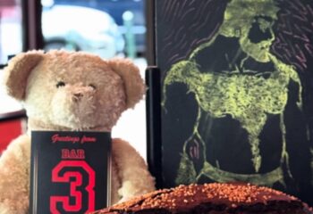 Bar TR3S Lisboa still life: Teddy bear holding a Greetings from Bar 3 card, next to a drawing of a muscular man on a chalkboard.