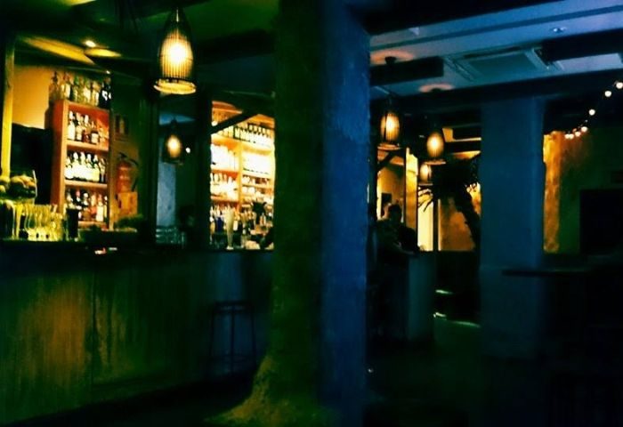 Baranoa Gay Bar interior in Madrid. Shelves of liquor behind bar. Dim lighting, tropical decor. Gay travel advice.