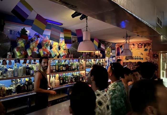 Inside Barbanarama Gay Bar in Madrid. Bartender, rainbow flags, and colorful busts above liquor bottles. Gay travel advice.