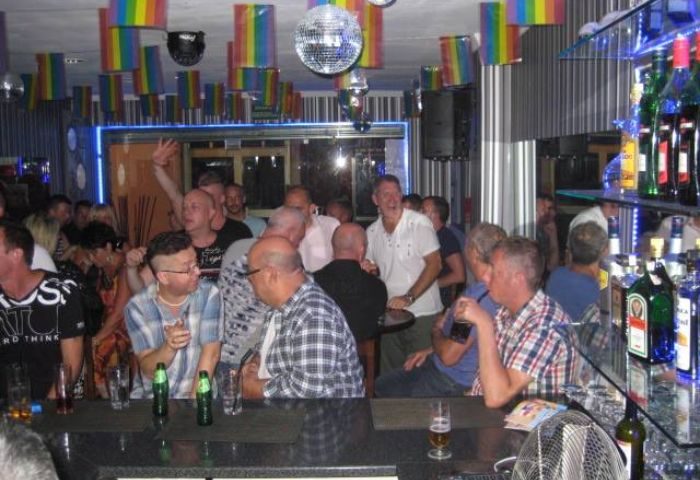 Inside Barcode Gay Bar in Benidorm, Spain. A lively scene with gay men enjoying drinks under rainbow flags and disco balls. Gay travel hotspot.