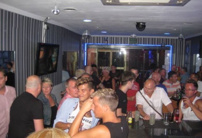 Inside Barcode Gay Bar in Benidorm, Spain. Crowded scene with men at the bar, disco balls, and blue neon lights. Gay travel.