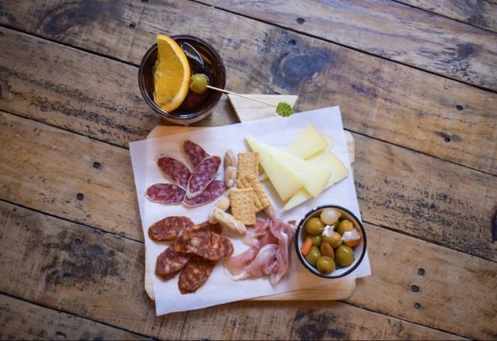 Bardaixa Gay Bar in Barcelona tapas: cheese, salami, crackers, olives, and ham served with a drink on a wooden table. Gay travel advice.