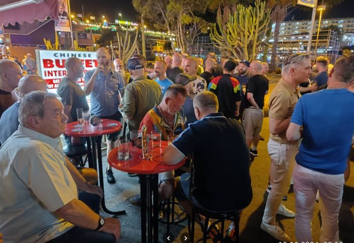 Bärenhöhle Bear Cave gay bar in Gran Canaria at night. A crowd of men gathers outside, enjoying drinks at tables. Gay travel destination.