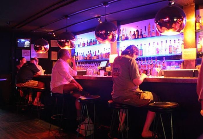 Interior of Barracuda Lounge, a gay bar in New York. Patrons sit at the bar, enjoying drinks under the ambient lighting.