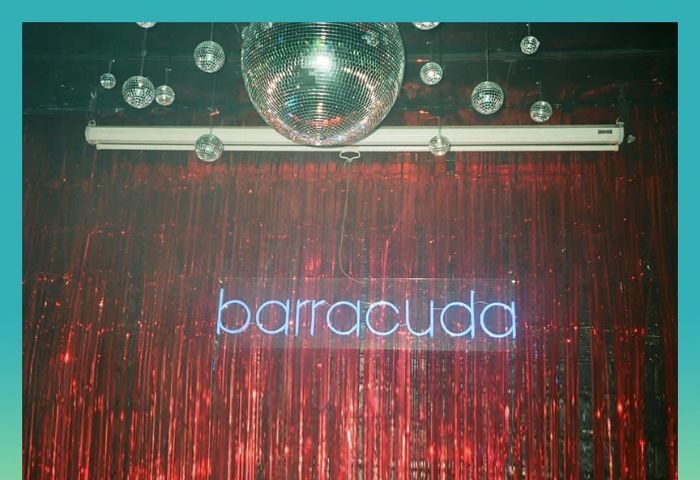 Barracuda Lounge, a gay bar in New York. Disco balls hang above a neon sign spelling barracuda against a red tinsel backdrop.