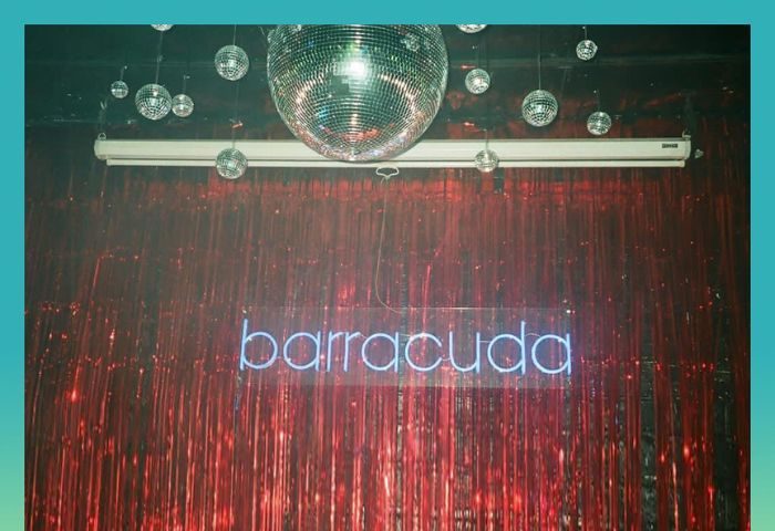 Barracuda Lounge, a gay bar in New York. Disco balls hang above a neon sign spelling barracuda against a red tinsel backdrop.