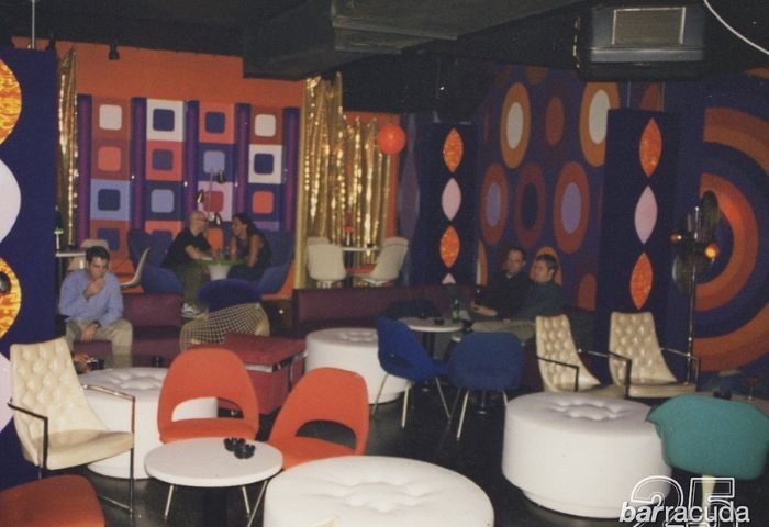 Interior of Barracuda Lounge, a gay bar in New York. People sit on colorful retro furniture with patterned walls in the background.