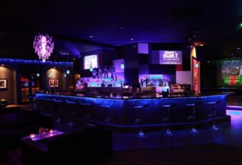 Bart Lounge dancefloor with blue lighting. Bar with stools, chandelier, and screens showing Street Fighter. Gay bar nightlife scene.