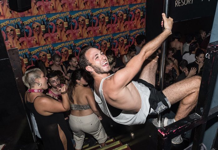 BeQueer: Man on pole at gay bar, Tropical Fever posters. Gay travel advice, nightlife. Queer scene, party atmosphere.