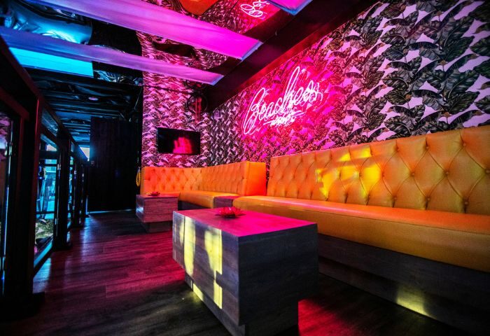 Interior of Beaches WeHo, a gay bar. Features yellow tufted banquettes, leaf-patterned wallpaper, neon sign, and colorful lighting. Gay travel advice.