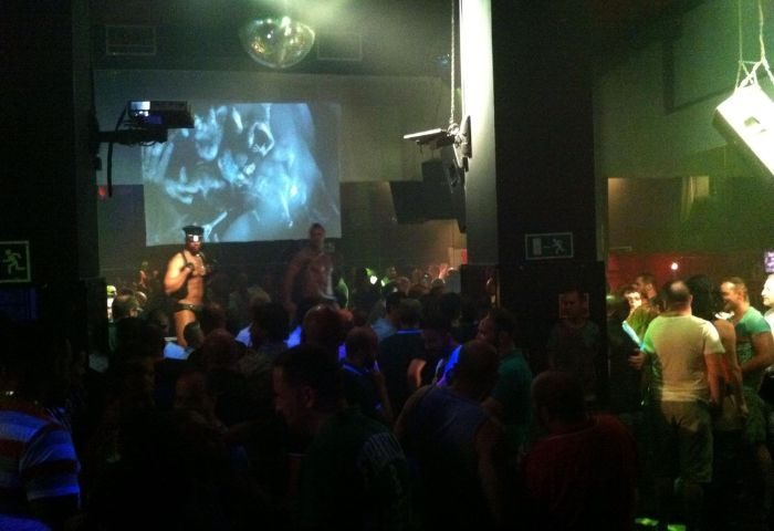 Crowded gay club in Madrid. A man in leather stands on stage. Disco ball, video projection, and green lighting. Bearbie gay club.