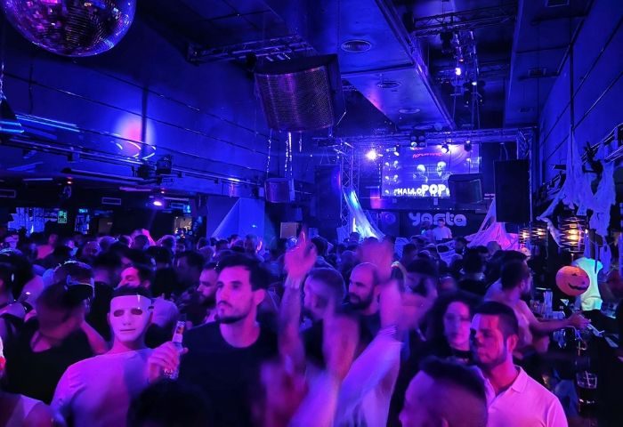 Crowded Bearbie gay club in Madrid. Men dance under blue lights, a disco ball, and stage. Halloween decorations visible.