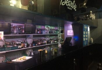 Bearbie Gay Club in Madrid: Bar area with liquor bottles on shelves, cutting board, and the club's logo visible. Gay travel.