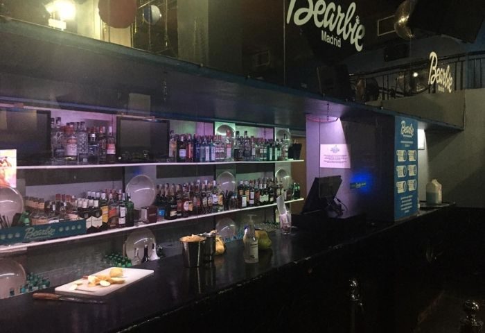 Bearbie Gay Club in Madrid: Bar area with liquor bottles on shelves, cutting board, and the club's logo visible. Gay travel.