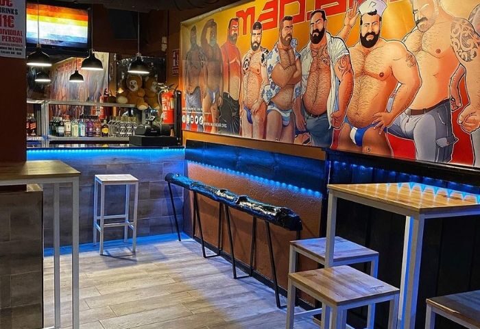 Interior view of Bears Bar, a gay bar in Madrid. Features a mural with bear men, blue lighting, and wooden tables/stools.