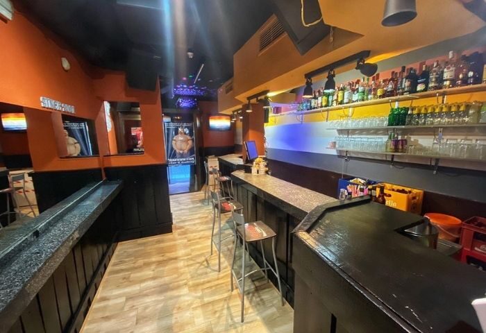 Interior of Bears Bar, a gay bar in Madrid, Spain. Bar stools line the counter. Shelves display bottles and glasses. Wood floors.