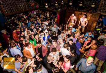 Crowded Beaux gay bar in San Francisco. Two shirtless dancers on stage. A diverse crowd of men and women partying. Nightlife scene.