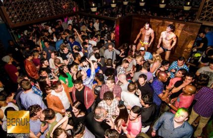 Crowded Beaux gay bar in San Francisco. Two shirtless dancers on stage. A diverse crowd of men and women partying. Nightlife scene.