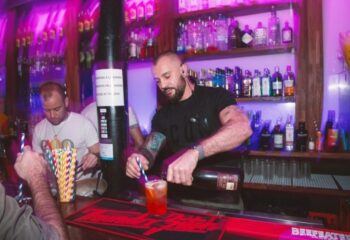 Bartender pours drink at Believe Club, a gay club in Barcelona, Spain. Nightlife scene with bar, bottles, and purple lighting.