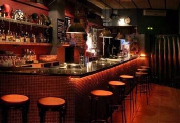 Interior of Berlin Dark gay cruise club in Barcelona. Bar with stools, shelves of bottles, and dim lighting. Gay travel advice.