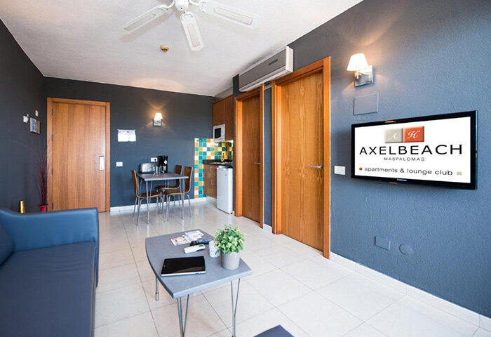 AxelBeach Maspalomas Apart & Lounge Club room. Adults-only gay men hotel interior with living area, kitchen, and AxelBeach TV logo.