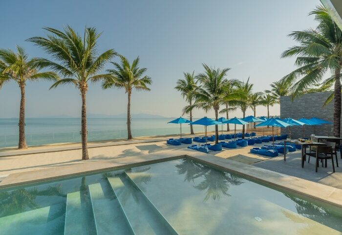 Explorar Koh Samui adult's only resort. Beachfront view with palm trees, blue umbrellas, and lounge chairs. Gay-friendly hotel and spa.