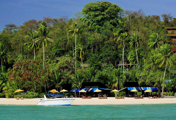Zeavola Hotel, Koh Phi Phi beachfront. Boat on turquoise water, sandy beach with umbrellas, tropical trees. Best gay-friendly hotel for gay men.