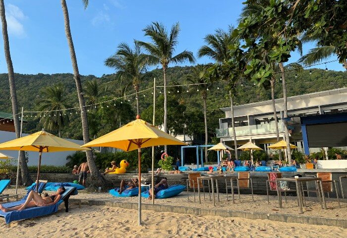 Explorar Koh Phangan Adults Only Resort beach view. Best gay hotels in Koh Phangan, Thailand. Beachfront lounge, palm trees, sunny day.