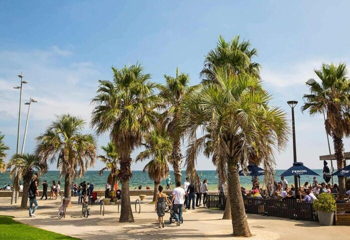 Saint Kilda Beach Hotel, a gay-friendly Melbourne spot. Palm trees line the beach where people stroll and relax at a beachfront cafe.