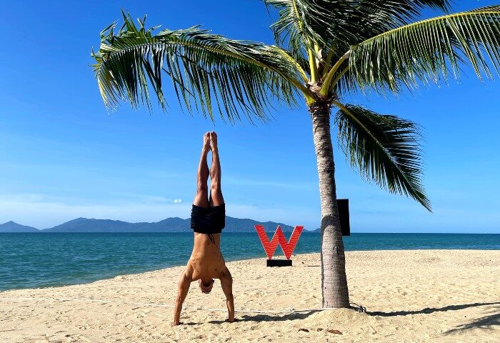 Gay man doing a handstand on the beach at W Koh Samui, a beachfront designed hotel. Palm tree and ocean view. Thailand travel.