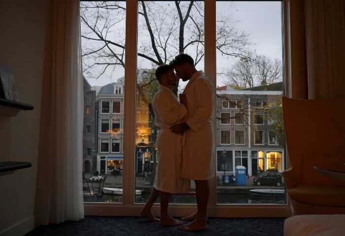 Gay honeymoon at Andaz Amsterdam Prinsengracht. A gay couple in robes embrace by a window overlooking a canal in Amsterdam, Netherlands.