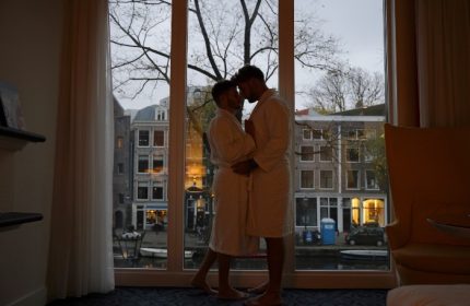 Gay honeymoon at Andaz Amsterdam Prinsengracht. A gay couple in robes embrace by a window overlooking a canal in Amsterdam, Netherlands.