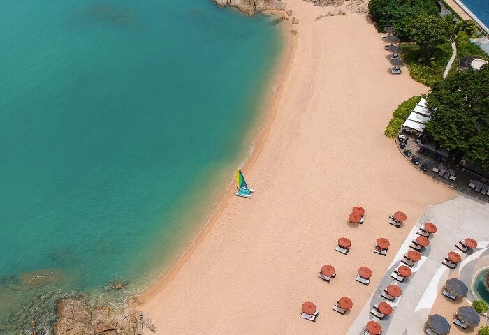 Garrya Tongsai Bay Samui private beach aerial view. Best gay honeymoon hotel in Koh Samui. Sailboat on the shore, umbrellas and beach chairs.