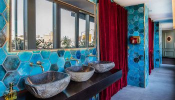 Bathroom at Axel Hotel Barcelona, a gay adults-only hotel. Stone sinks, blue hexagon tiles, and red velvet curtains create a stylish urban spa.
