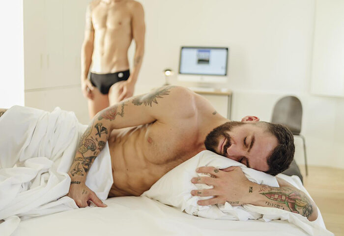 The Late Birds Lisbon Gay Urban Resort: Man sleeping in bed, tattooed, with another man standing in the background. Gay men only hotel in Lisbon.