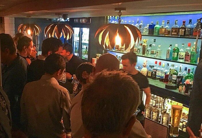 New Bloomsbury Set, a gay bar in London. Crowd of patrons at the bar with a bartender. Shelves of liquor bottles behind the bar.