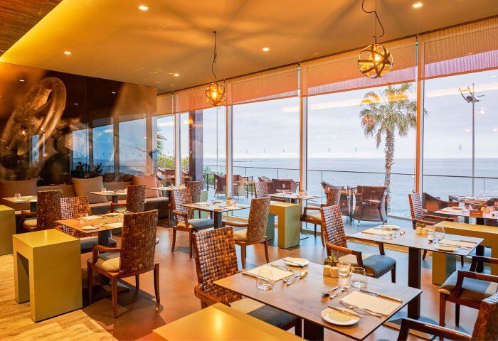 Saccharum Hotel Madeira restaurant interior. Beach spa resort, gay-friendly hotel, ocean view, tables, chairs, and palm tree. Portugal.
