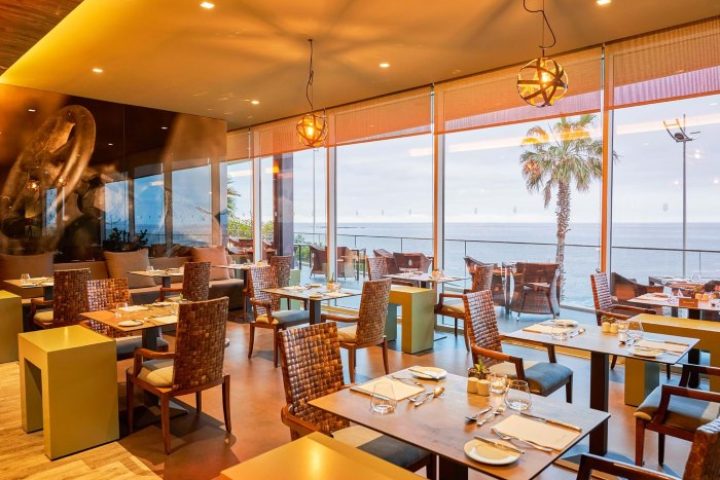 Saccharum Hotel Madeira restaurant interior. Beach spa resort, gay-friendly hotel, ocean view, tables, chairs, and palm tree. Portugal.