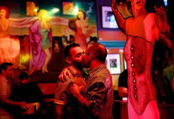 Gay bar in Chicago: Two men kissing at a big chicks fetish bar. Red lighting, mural of women in the background, and a corset display.