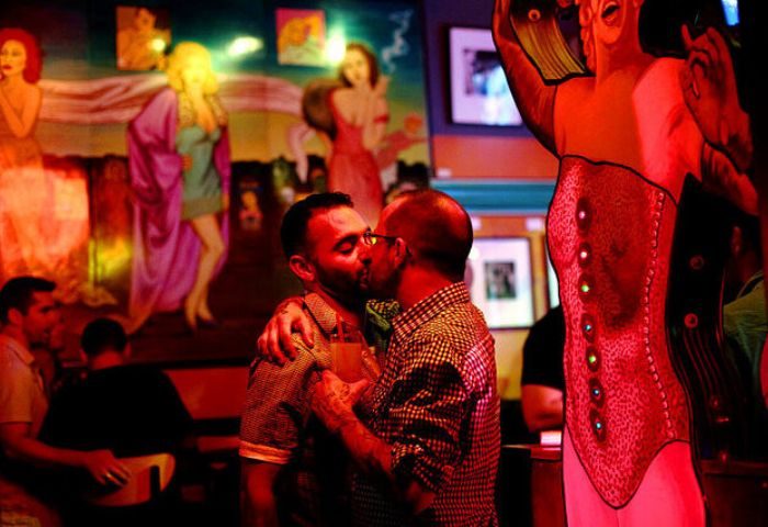 Gay bar in Chicago: Two men kissing at a big chicks fetish bar. Red lighting, mural of women in the background, and a corset display.
