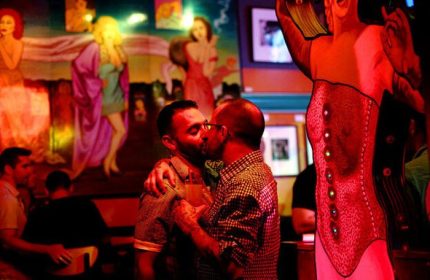 Gay bar in Chicago: Two men kissing at a big chicks fetish bar. Red lighting, mural of women in the background, and a corset display.