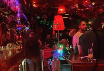 Big Bar interior. Gay bar scene with men at the bar, red lighting, and festive decorations. Nightlife and travel advice.