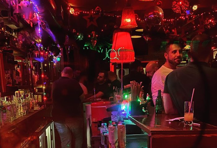 Big Bar interior. Gay bar scene with men at the bar, red lighting, and festive decorations. Nightlife and travel advice.